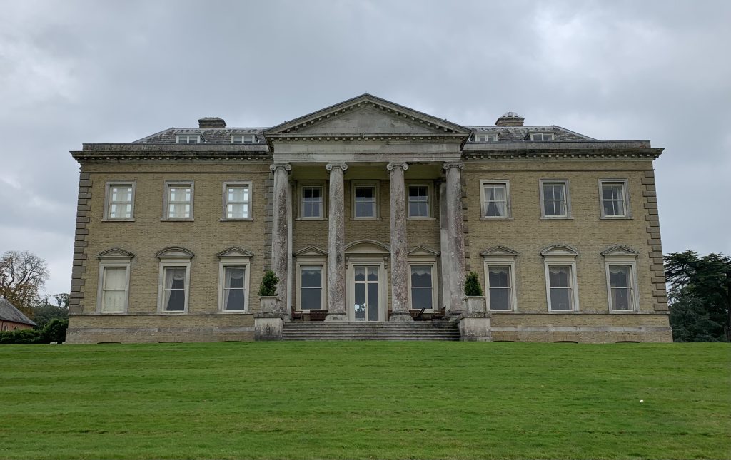 a large brick building with green grass in front of a house with Basildon Park in the background