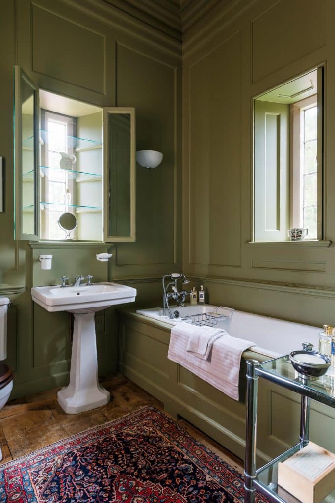 Olive green panelled bathroom with glass shelving in window alcove and antique rug